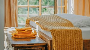 A bed with a white sheet and a yellow blanket, a wooden nightstand with folded towels, and a window with curtains. - Living among orchards by Lake Chiemsee (Übersee)