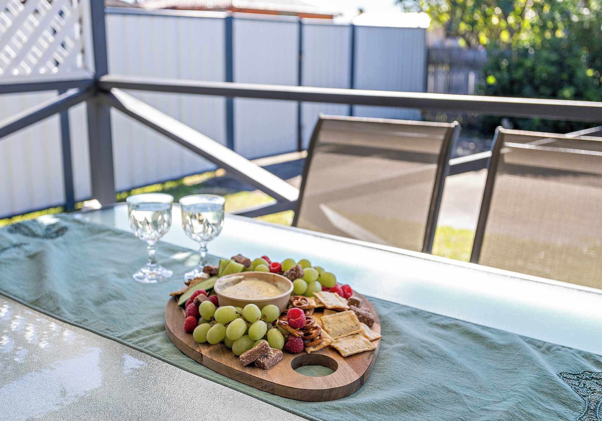 Una mesa con cubierta de vidrio, una tabla de madera con uvas, nueces y galletas, y dos copas de vino.