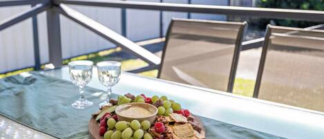 Una mesa con cubierta de vidrio, una tabla de madera con uvas, nueces y galletas, y dos copas de vino.