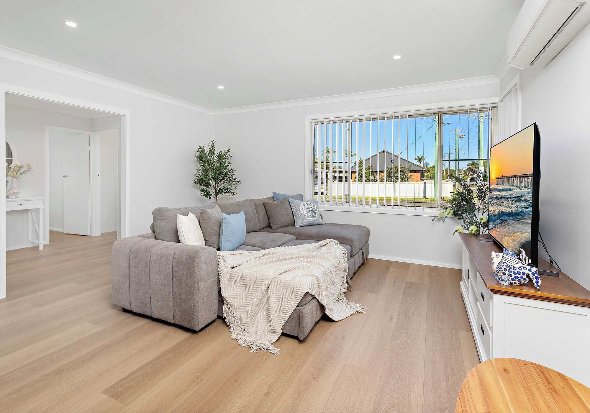 A modern living room with a grey sofa, a TV on a wooden stand, and a large window with blinds.