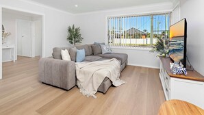 Una sala de estar moderna con un sofá gris, un televisor sobre un mueble de madera y un ventanal con persianas.