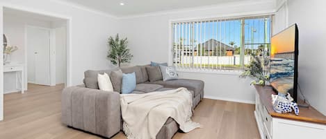 Een moderne woonkamer met een grijze bank, een tv op een houten dressoir, en een groot raam met zonwering.
