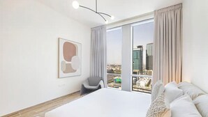 Ett modernt hotellrum med en stor säng, en stol, utsikt över staden och ett inramat konstverk på väggen.