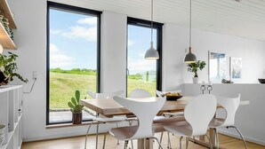A modern kitchen with white cabinetry, a wooden dining table, and chairs. There are large windows offering a view of a green landscape. - Summerhouse close to nature by The Limfjord (Nykøbing Mors)