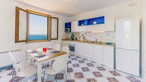 A compact kitchen with white cabinets, a microwave, and a refrigerator. There's a dining table with chairs and a view of the sea through a window. - Baroque house with lemon groves in Amalfi (Amalfi)