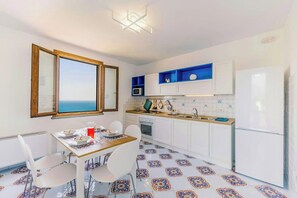 Eine kompakte Küche mit weißen Schränken, einer Mikrowelle und einem Kühlschrank. Ein Esstisch mit Stühlen und Blick aufs Meer durch ein Fenster.