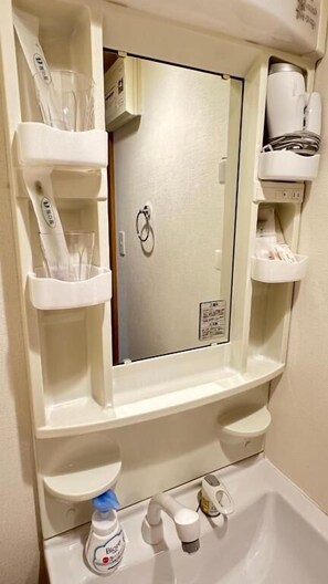 A bathroom with a mirror, shelves for toiletries, a sink, and a shower. - Dc Sakura Hotel - daikokuyon Branch (Osaka)