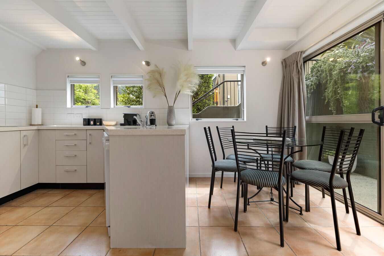 A modern kitchen with a central island, a dining area with chairs, and large windows offering a view of greenery.