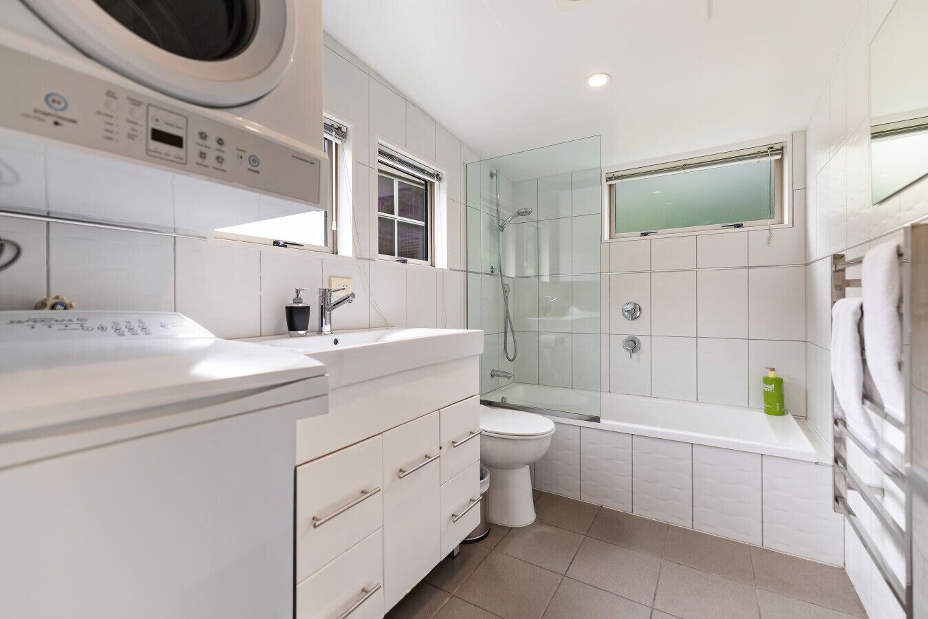 Une salle de bain moderne dotée d’un lave-linge intégré, d’une douche à parois de verre et d’un sol carrelé de couleur blanche.