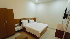 A hotel room with a bed, a wooden wardrobe, a nightstand with a bottle, a small ottoman, and a television mounted on the wall.