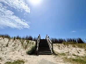 Eine Holztreppe führt über einen sandigen Strand mit Dünen und Gras hinauf.