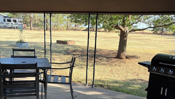 An outdoor patio with a table and chairs, a grill, and a view of a grassy field and trees.