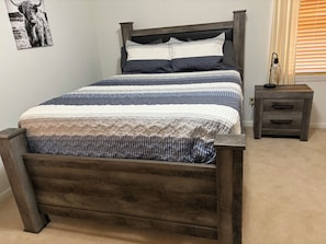 A bed with a wooden frame, a headboard, and two pillows. There is a bedside table with drawers and a lamp.
