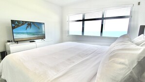Ein Schlafzimmer mit einem Bett, einem Fernseher auf einem Schrank, einem Fenster mit Jalousien und Blick aufs Meer.