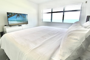 A bedroom with a bed, a TV on a stand, a window with blinds, and a view of the sea.