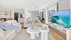 A modern living room with a large flat-screen TV, a sofa, a coffee table, and a dining area. - Drift n Dream Apartment w/Free Parking+Pool+Gym (Surfers Paradise)