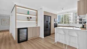 A modern kitchen with a built-in refrigerator, a compact microwave, and a small coffee machine. There is a white countertop with a sink and bar stools. - Drift n Dream Apartment w/Free Parking+Pool+Gym (Surfers Paradise)