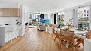 A modern kitchen with white cabinetry, a dining area with a round table and chairs, and a large window with a view of buildings and greenery. - Drift n Dream Apartment w/Free Parking+Pool+Gym (Surfers Paradise)