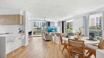 A modern kitchen with white cabinetry, a dining area with a round table and chairs, and a large window with a view of buildings and greenery.