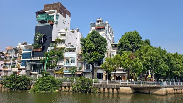 Vista do rio de um horizonte urbano com edifícios modernos, uma ponte para pedestres e vegetação exuberante.