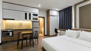 Quarto de hotel compacto com cozinha americana, uma cama, escrivaninha e uma cadeira.