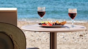A table with a plate of food and two glasses of red wine, set against a beach backdrop with the sea and a clear sky. - Fodele Beach Front Apts. (Malevizi)
