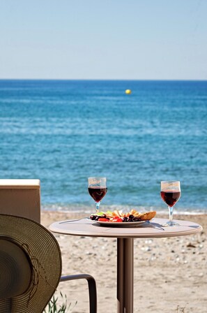 Ein Tisch mit einem Teller Essen und zwei Gläsern Rotwein, vor einer Strandkulisse mit Meer und blauem Himmel.