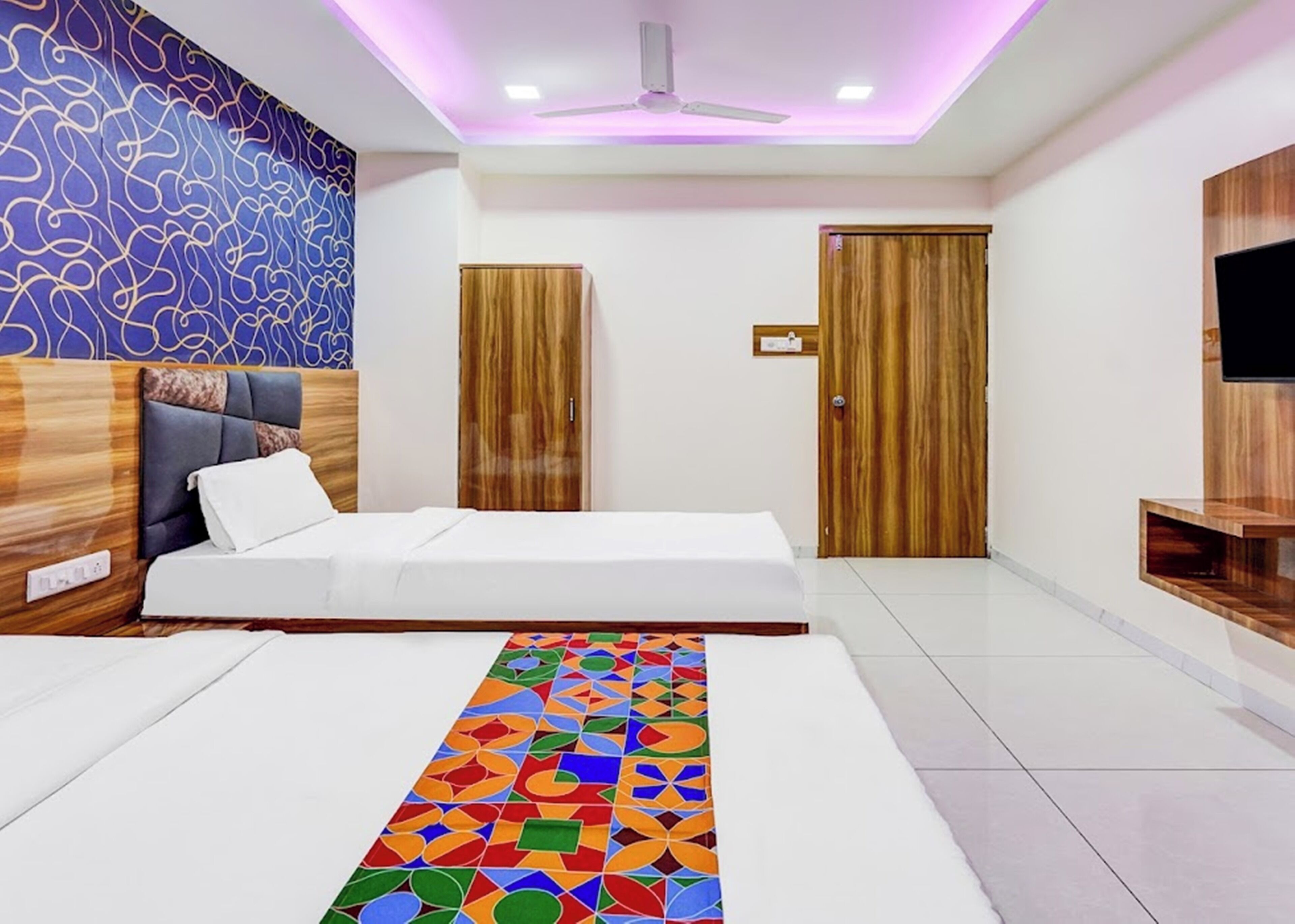 Una camera d'albergo moderna con un letto, una ventola a soffitto, un televisore fissato al muro e un tappeto decorato.