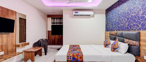 A hotel room with a bed, a desk, a chair, a TV, and an air conditioning unit.