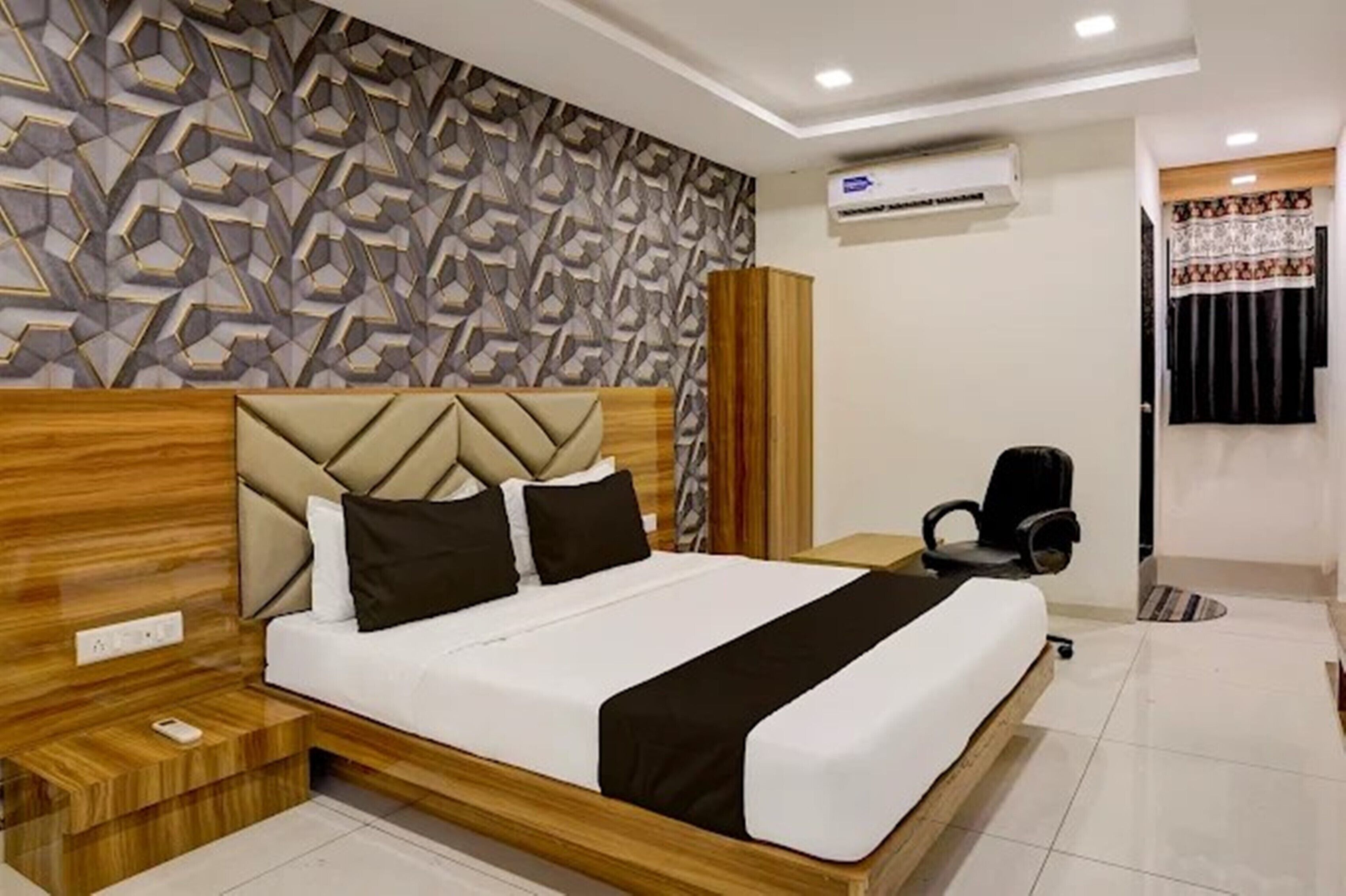 Ein modernes Hotelzimmer mit Wand im geometrischen Muster, einem hölzernen Kopfteil, einem Bett mit weißer und schwarzer Bettwäsche, einem Schreibtisch mit Stuhl und einer Klimaanlage.