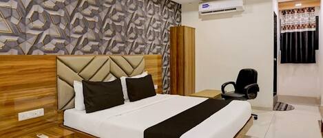 A modern hotel room with a geometric-patterned wall, a wooden headboard, a bed with white and black bedding, a desk with a chair, and an air conditioning unit.
