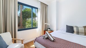 Habitación de hotel con un ventanal grande, una cama con ropa de cama estampada, una silla y una mesita de noche con una lámpara y una planta.