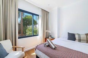 A hotel room with a large window, a bed with patterned bedding, a chair, and a nightstand with a lamp and a plant. - Resort Escape: King Bed, Pool & Balcony (Darwin City)