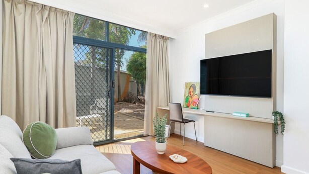 Una sala moderna con un sofá, una mesa de centro, un televisor montado en la pared y un ventanal corredizo que da a un área exterior.