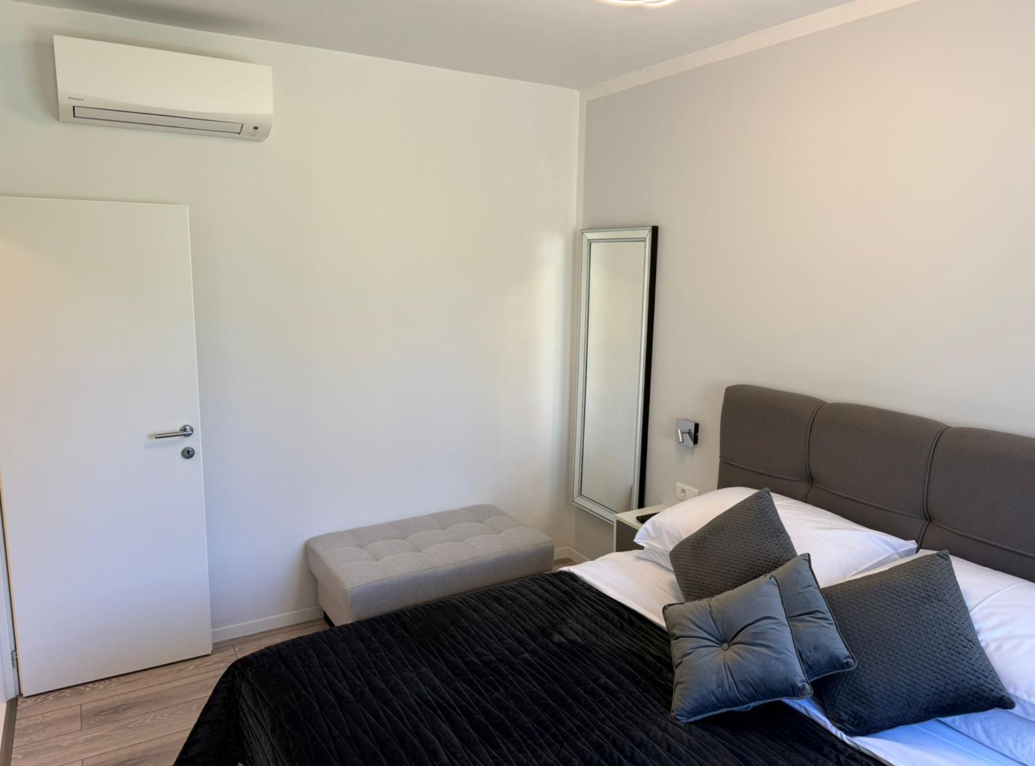 A hotel room with a bed, a bench, a large mirror, and an air conditioning unit.