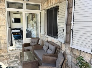 Une terrasse couverte, aménagée avec des meubles en osier, une table en verre et offrant une vue sur l’intérieur.