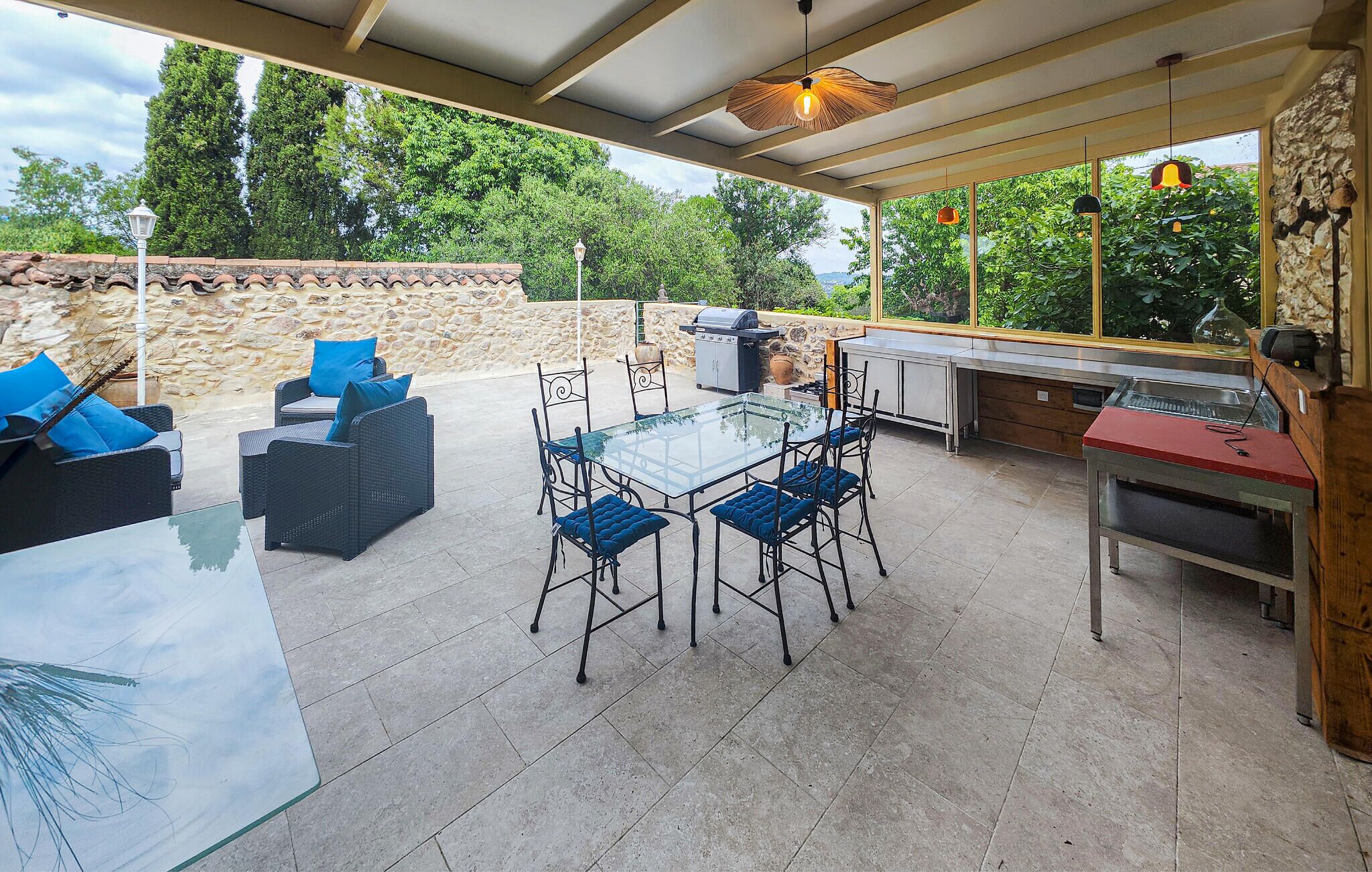 An outdoor patio with a glass table, blue cushioned chairs, a grill, and a stone wall.