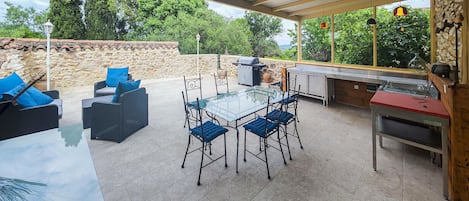 An outdoor patio with a glass table, blue cushioned chairs, a grill, and a stone wall.