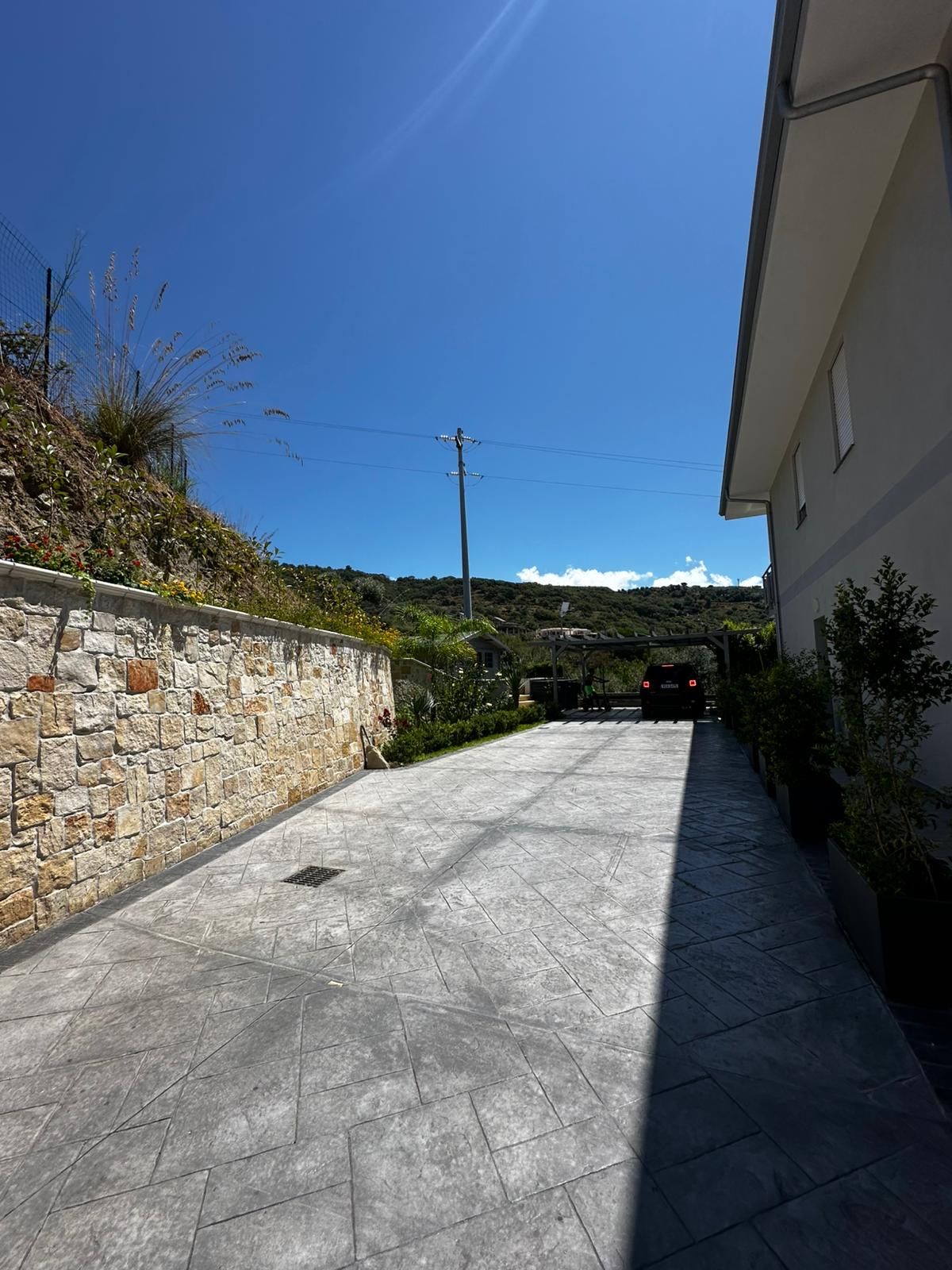 Un chemin pavé mène à un bâtiment dont un côté est bordé d’un mur de pierre et l’autre d’une clôture en métal.