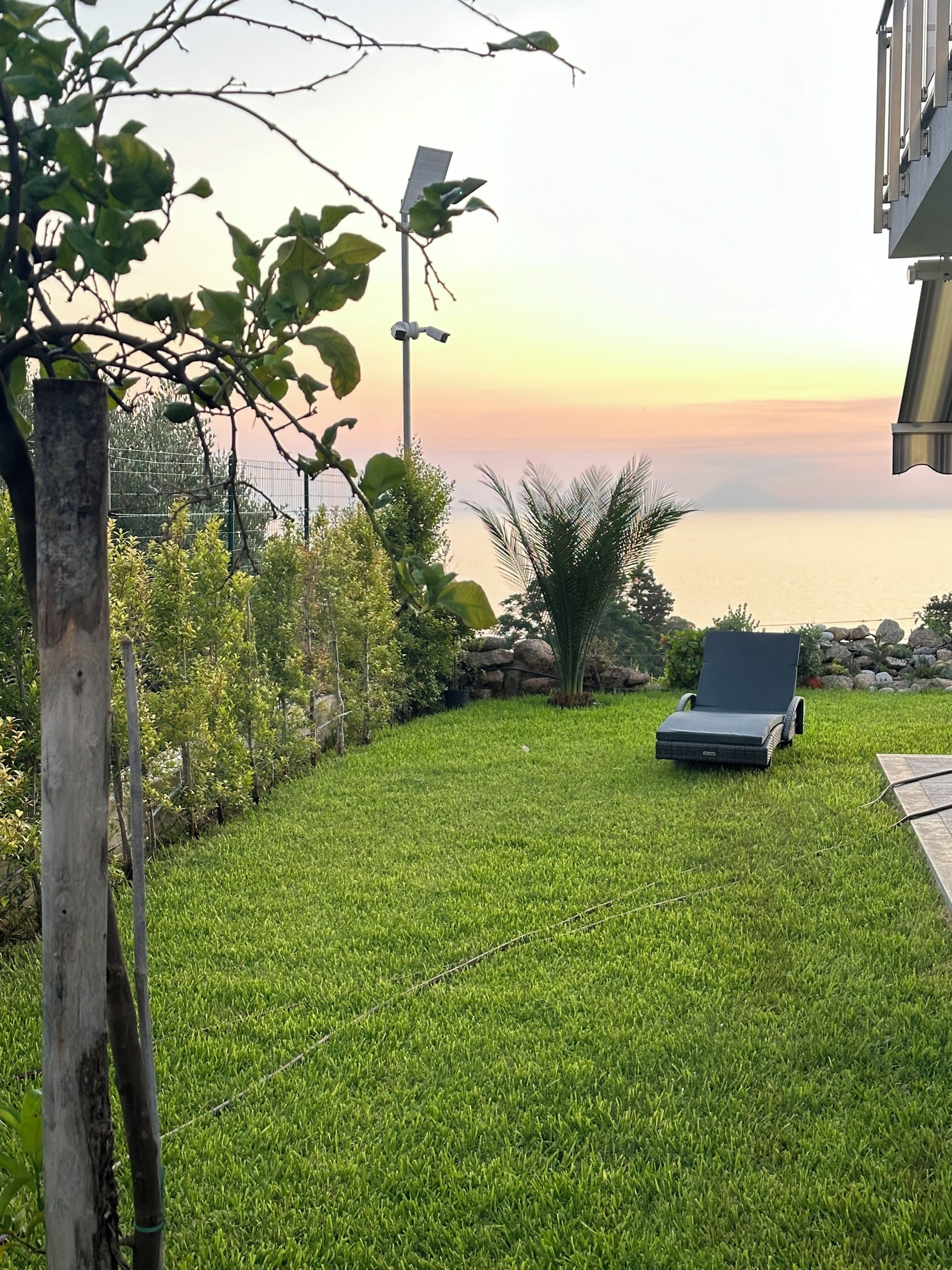 A grassy lawn with a lounge chair, a solar-powered street lamp, and a view of the sea at sunset.