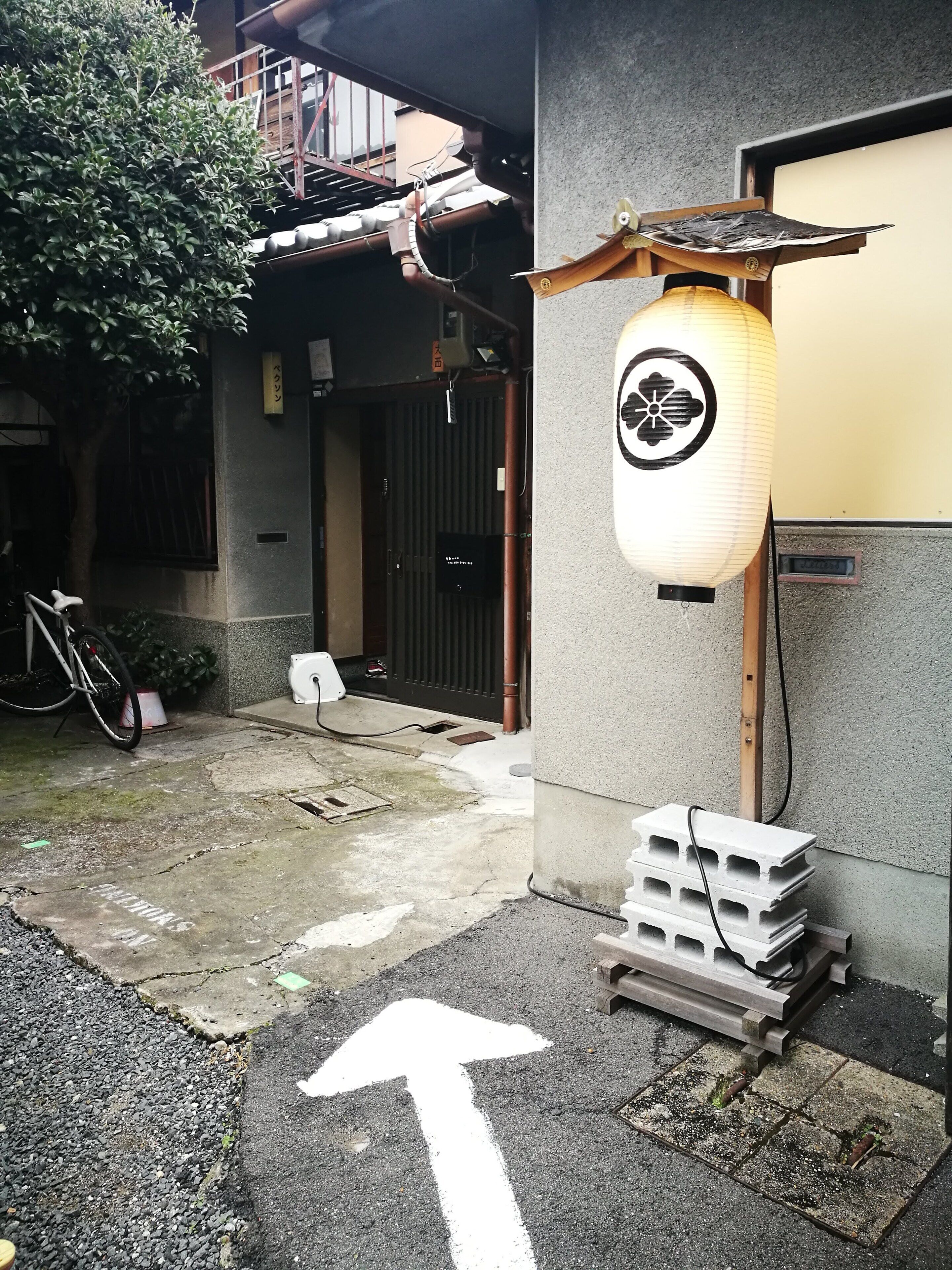 A building entrance with a traditional Japanese lantern, a bicycle, and a directional arrow on the ground.
