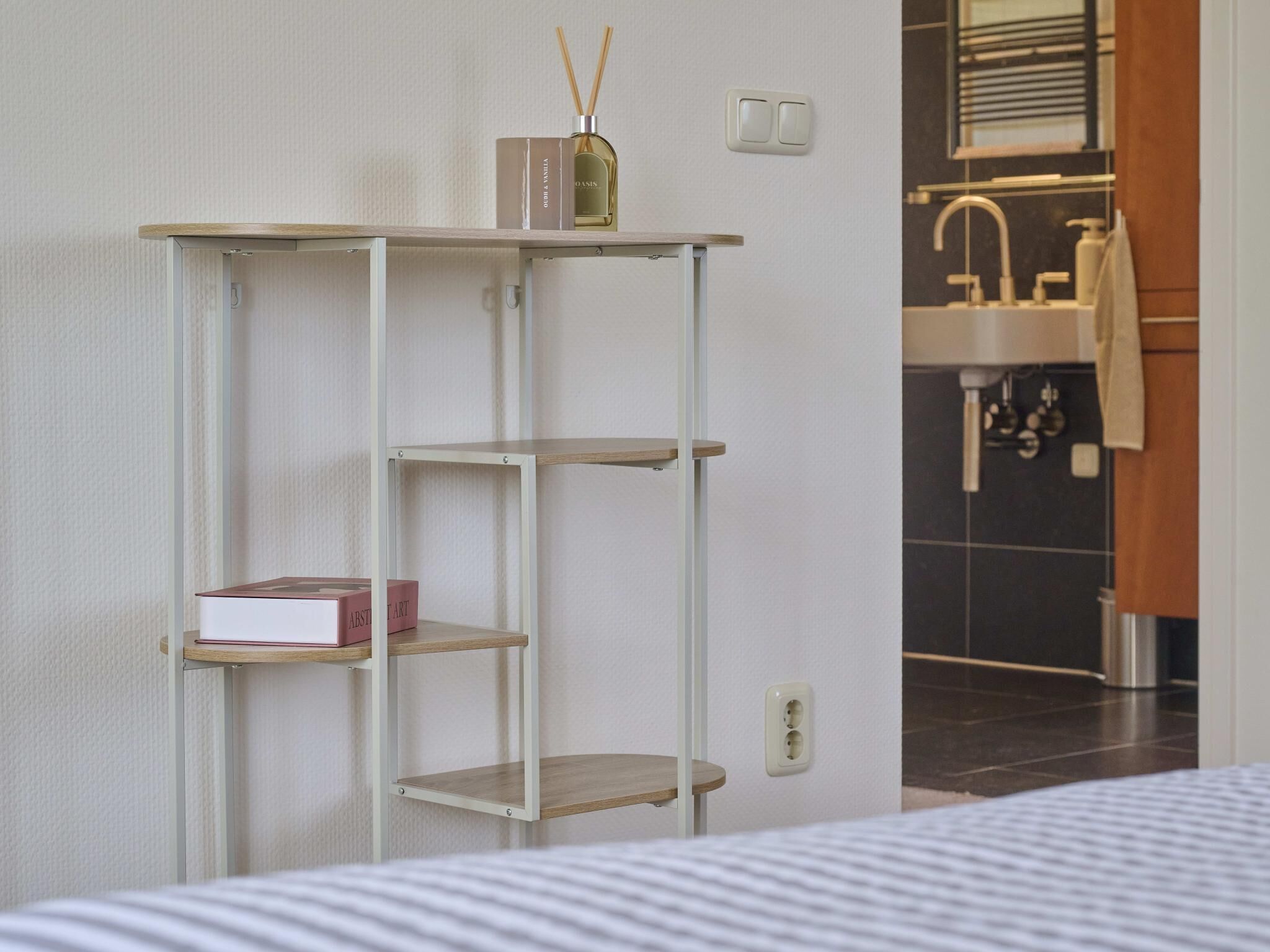 Een kamer met een bed, een witte muur, een plank met boeken en een fles, en een badkamer zichtbaar door een geopende deur.