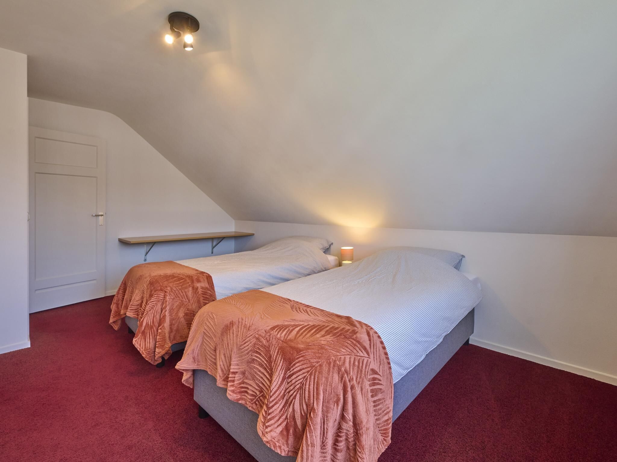 A room with two beds, a sloped ceiling, a small shelf, and a door.