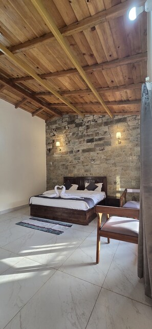Quarto com teto de madeira, parede de tijolos, cama com roupa de cama branca, duas luminárias de cabeceira e uma cadeira de madeira.