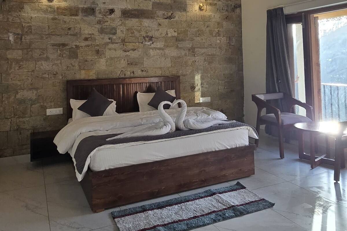 Un dormitorio con pared de piedra, vigas de madera en el techo, cama con ropa blanca y vista por una ventana.