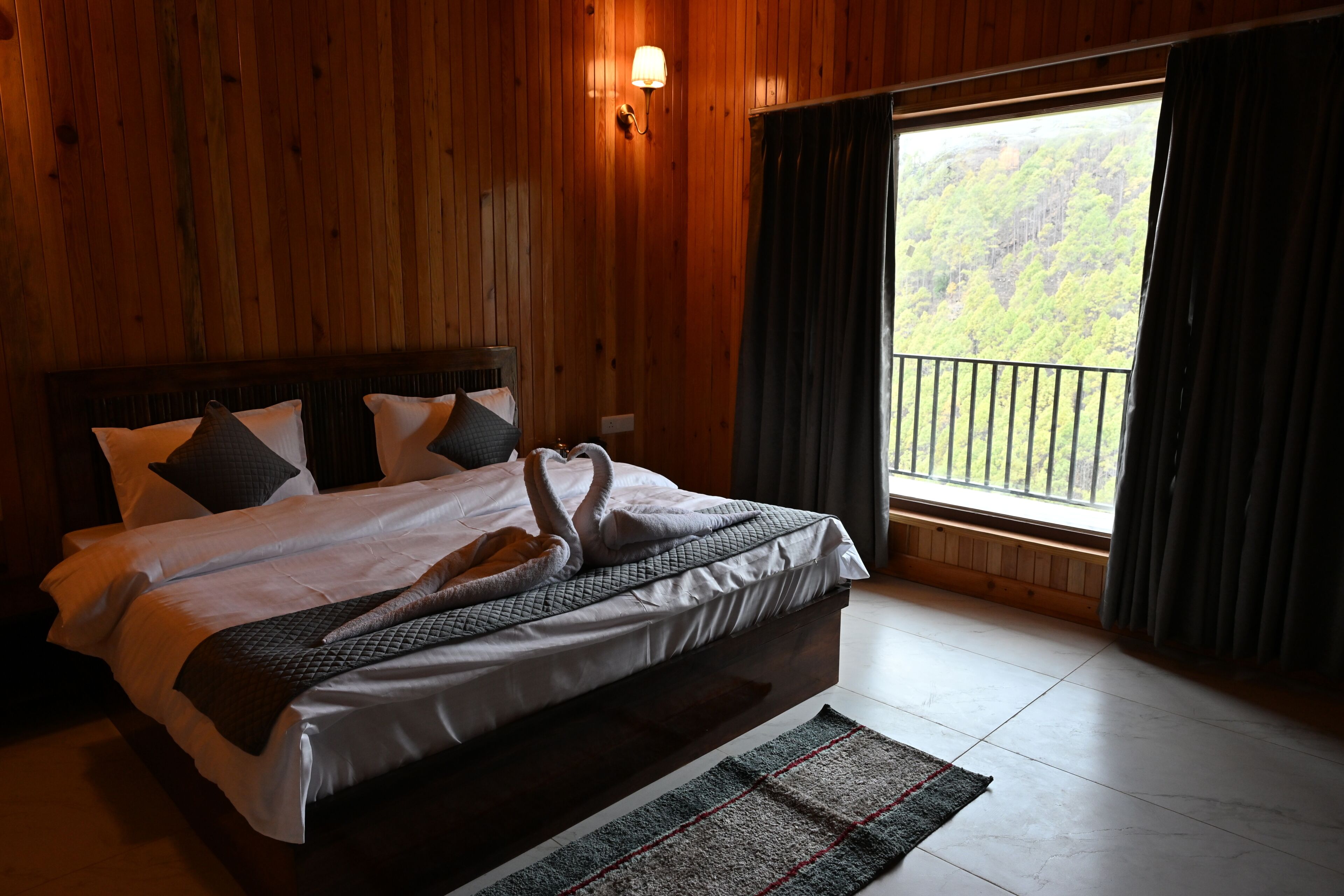 A hotel room with a wooden interior, a large bed with a white and gray duvet, a headboard, and a view of a forest through a window.