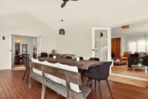 A dining area with a wooden table, benches, and chairs, a ceiling fan, and a view into another room with a sofa and a window.