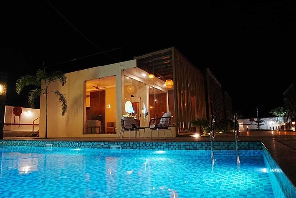 A modern building with a pool, a dining area with chairs, and a visible entrance.