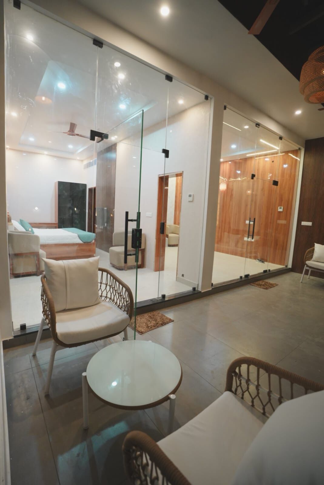 Un intérieur moderne avec des murs en verre, un coin salon doté de fauteuils en osier et d’une table ronde, et une chambre visible avec un lit et un grand miroir.