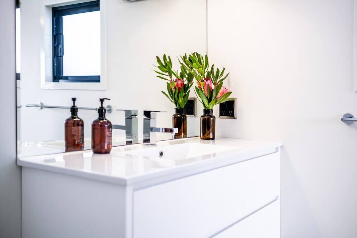 A modern bathroom with a white vanity, two bottles of liquid soap, and two vases with green plants.